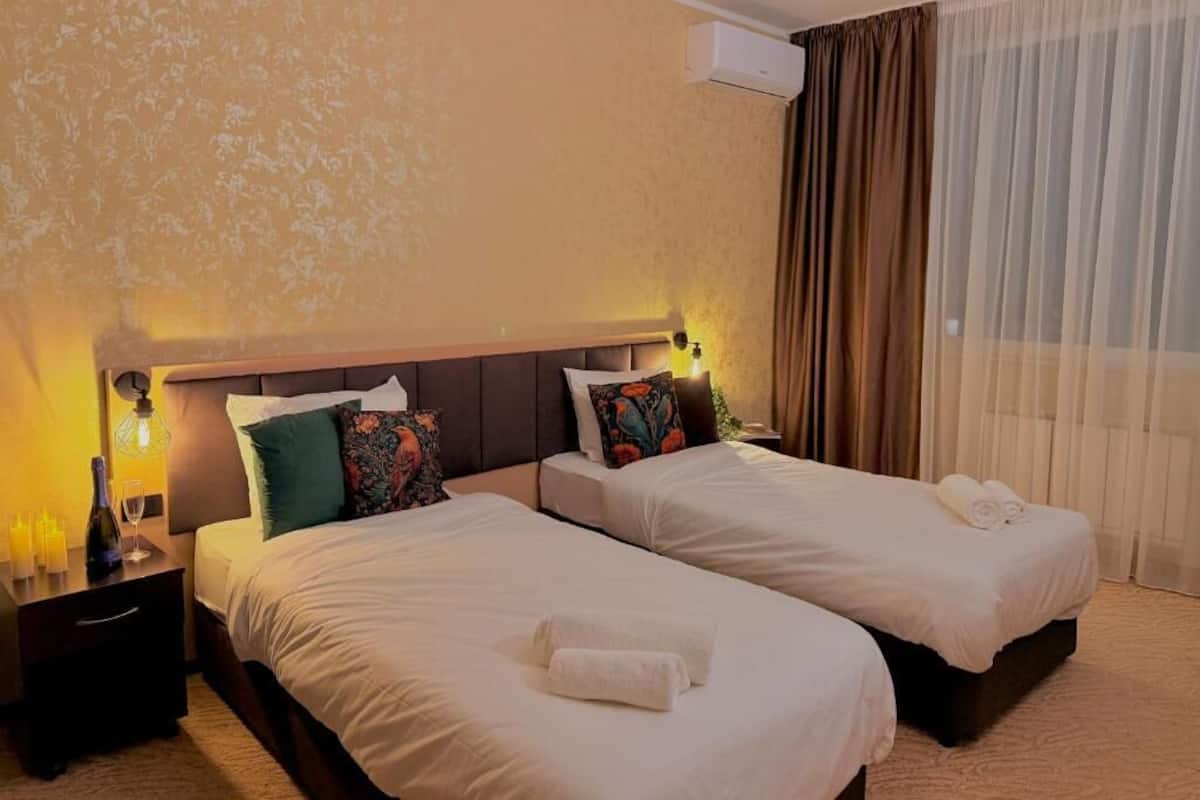 Habitación de hotel con dos camas, una mesita de noche con lámpara, aire acondicionado en la pared y una ventana con cortinas.