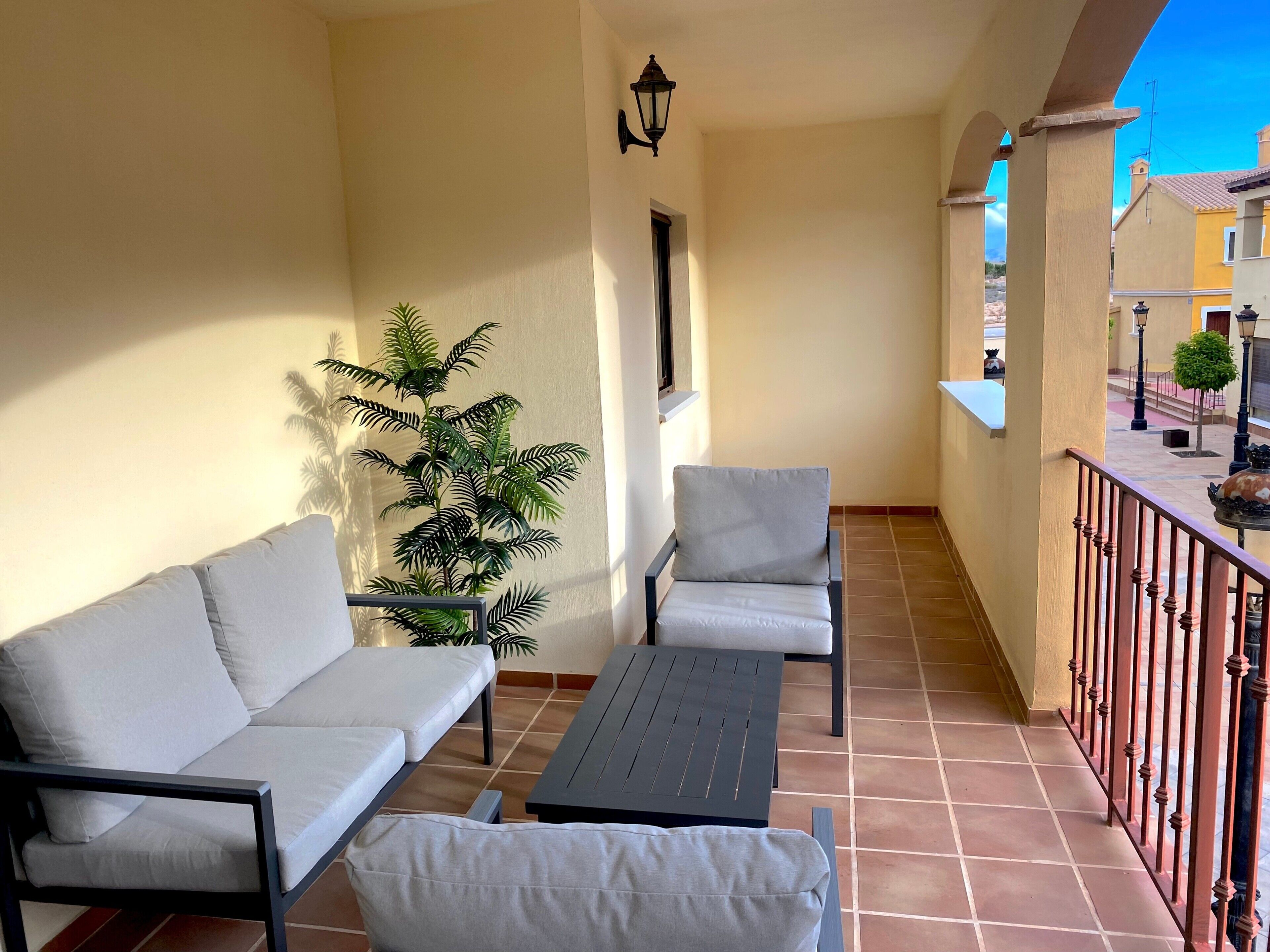 A balcony with a sofa, chairs, a table, a potted plant, and a railing.