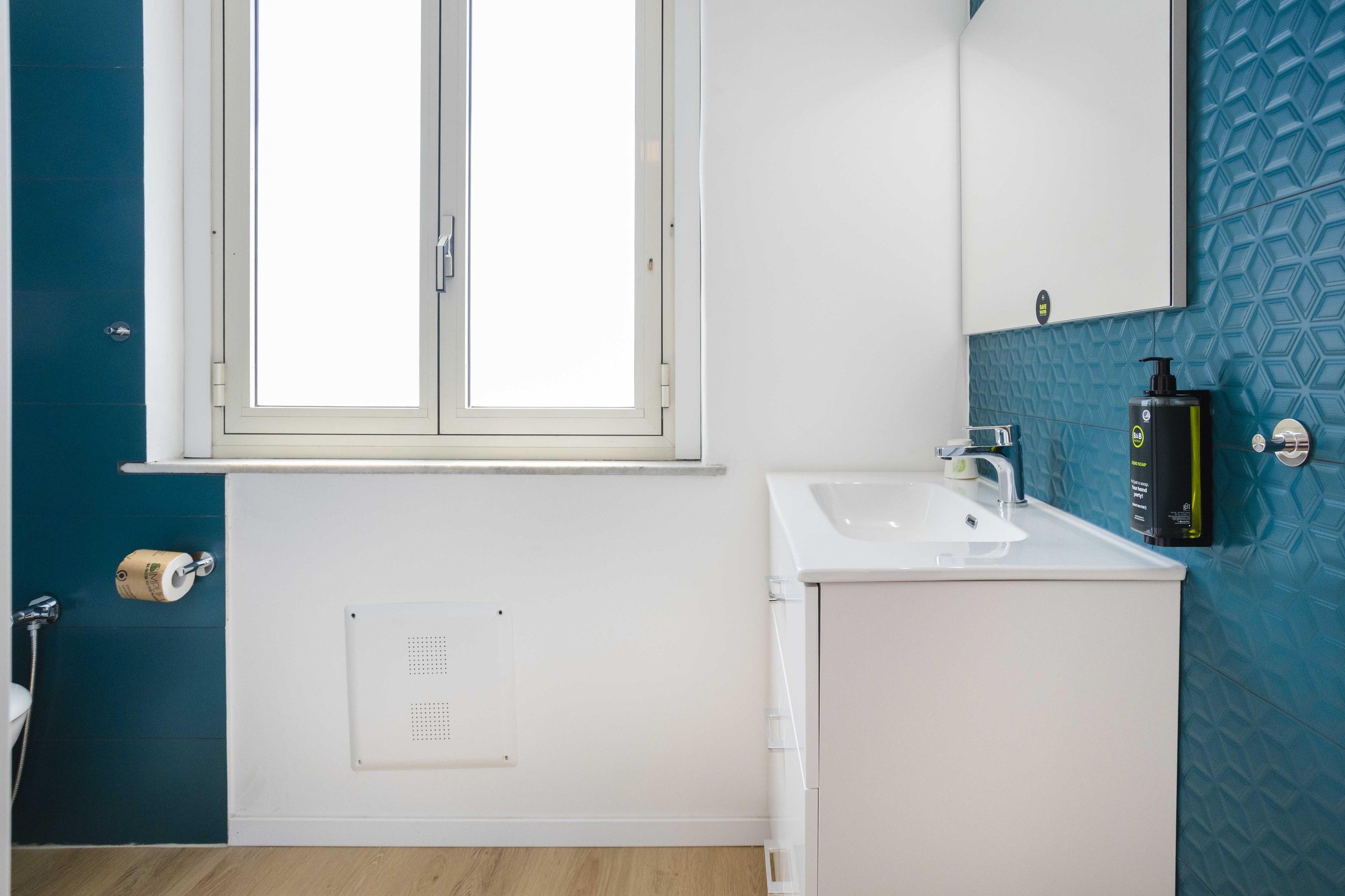 Un baño con un lavamanos blanco, un espejo encima y una ventana con marco blanco.
