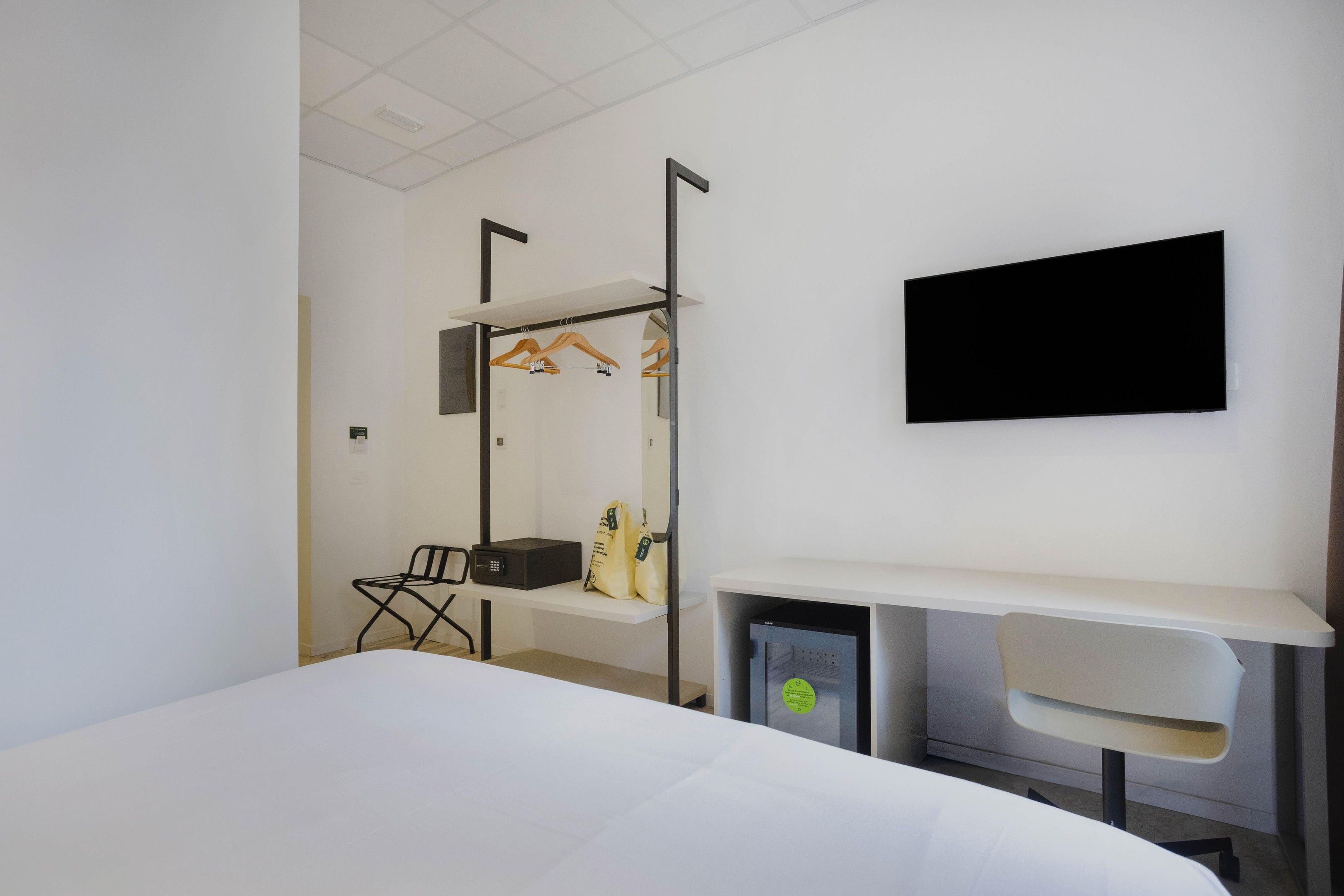 Habitación de hotel con cama, escritorio, silla, televisor montado en la pared y perchero con perchas.