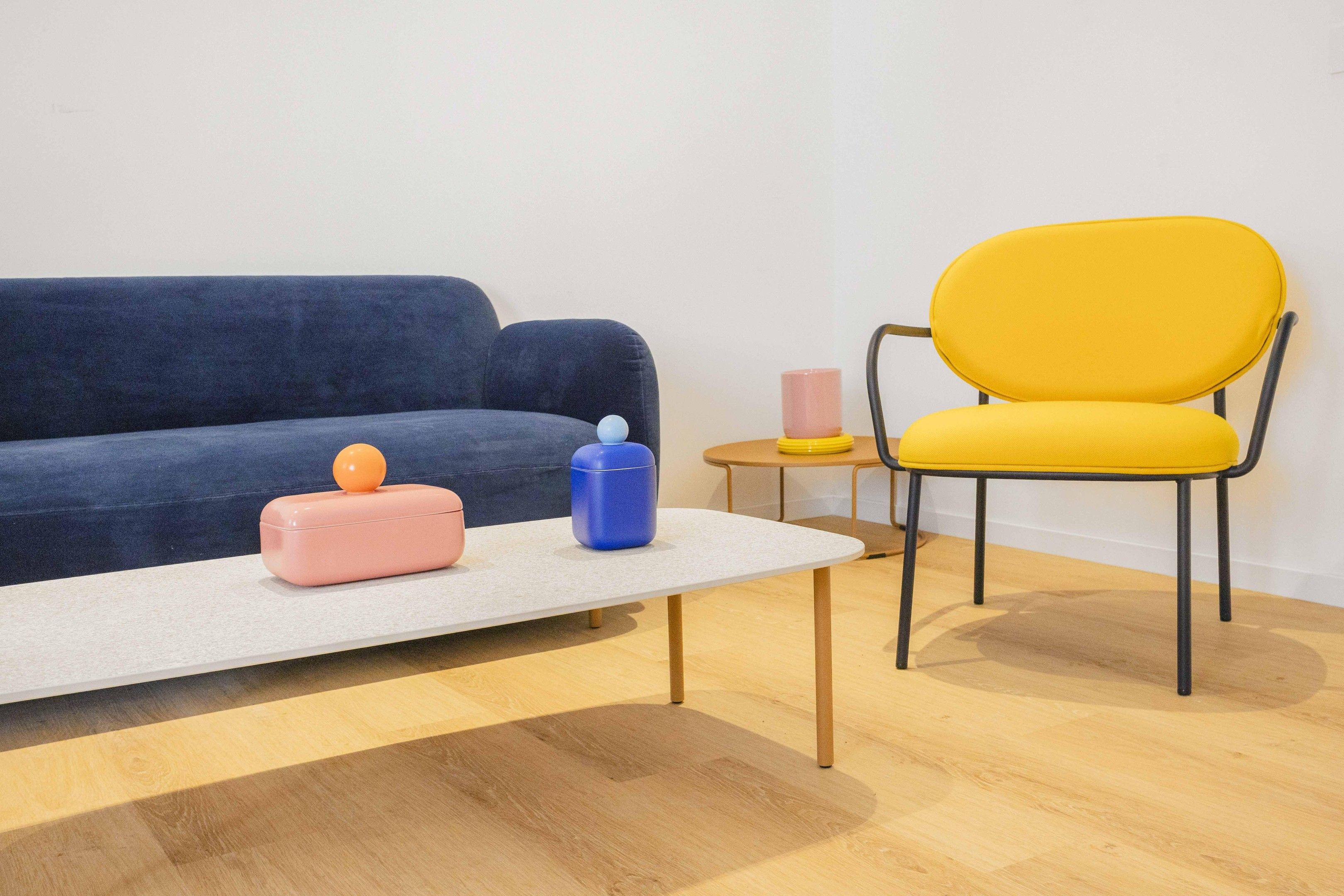 A room with a blue sofa, a yellow chair, a small round table with a pink mug, and two containers on a white table.
