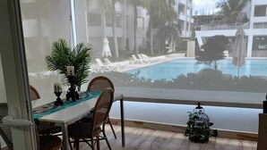 Een balkon met een eettafel, stoelen, een potplant en uitzicht op een zwembad en gebouwen.