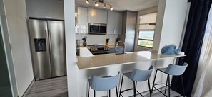 Una cocina moderna con un refrigerador de acero inoxidable, un microondas y un lavamanos. Hay tres taburetes azules y una ventana con vistas.
