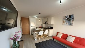 Una sala moderna con un sofá rojo, un comedor con mesa y sillas, una cocina con refrigerador y microondas, y un pasillo que lleva a una puerta.