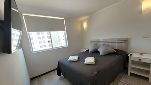 Una habitación de hotel moderna con una cama grande, una mesita de noche, un televisor de pantalla plana en la pared y una ventana con vistas a edificios.
