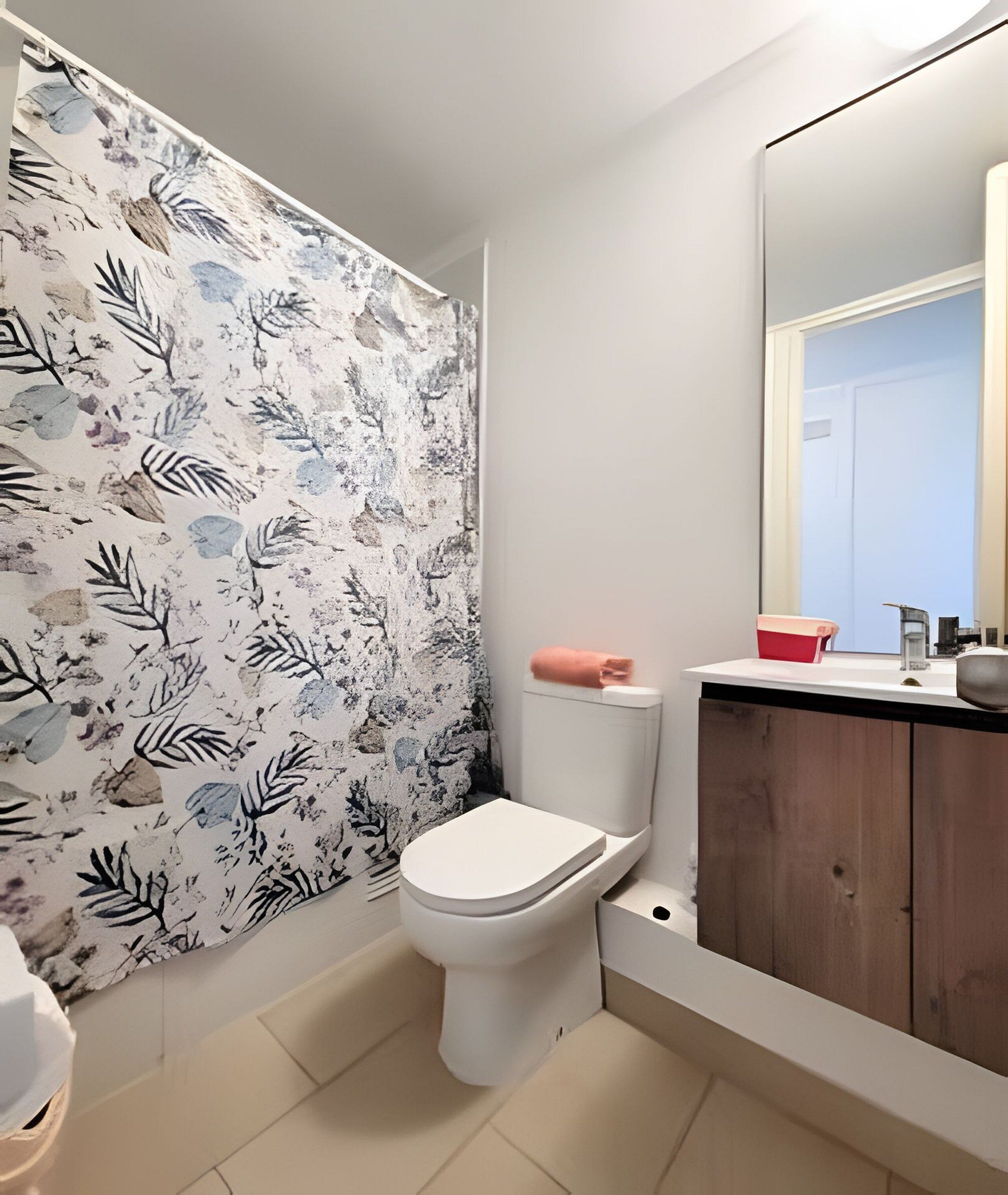 A bathroom with a toilet, a shower curtain with leaf patterns, a wooden cabinet, and a mirror.