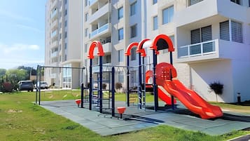 Um parquinho infantil com escorrega e estruturas de escalada em frente a um prédio de apartamentos moderno.
