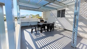 Ett takförsett utomhusområde med ett bord och stolar, en inbyggd grill och utsikt över det omgivande landskapet.