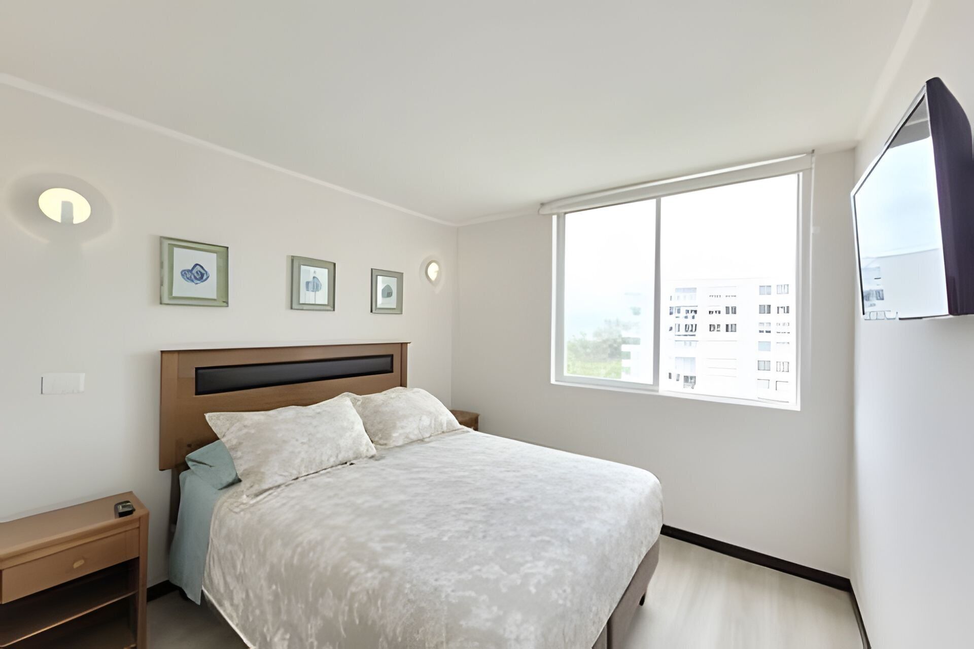 Un dormitorio con una cama, una mesita de noche, un televisor y una ventana con vistas a edificios.