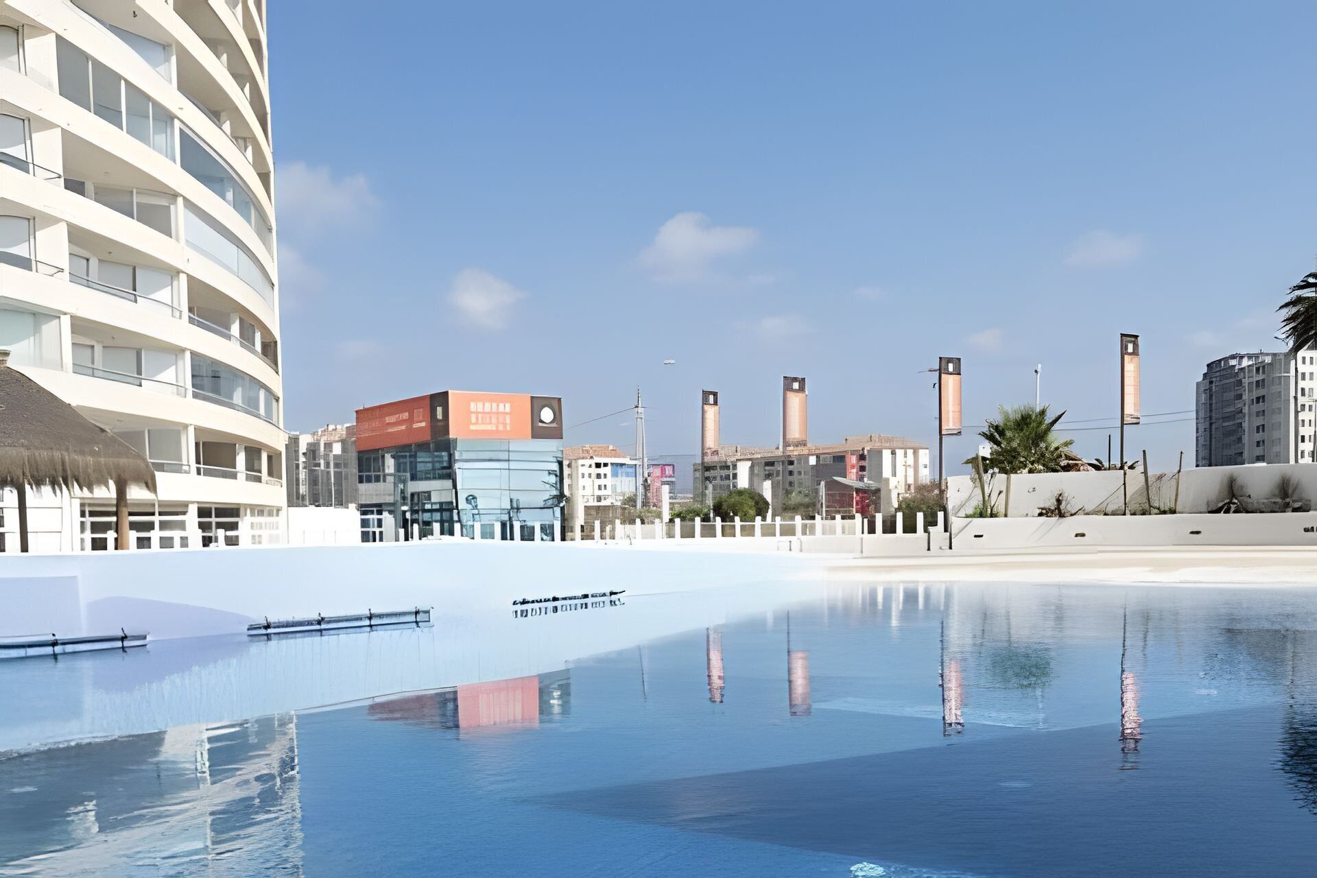 A modern hotel with a pool, a thatched-roof structure, and a clear blue sky.
