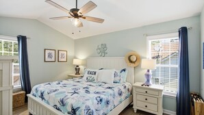 A bedroom with a bed, a ceiling fan, a nightstand, a lamp, a window with curtains, and a wall with framed pictures. - Tracie's Oasis - Pool/fenced/beach walkable! (Oak Island)