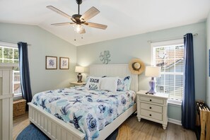 Ein Schlafzimmer mit Bett, Deckenventilator, Nachttisch, Lampe, Fenster mit Vorhängen und einer Wand mit gerahmten Bildern.