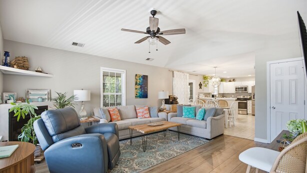 A spacious living room with a ceiling fan, a sofa, a coffee table, and a kitchen in the background. - Tracie's Oasis - Pool/fenced/beach walkable! (Oak Island)