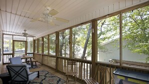 A covered porch with wooden flooring, a ceiling fan, and large windows offering a view of trees and a house. - Tracie's Oasis - Pool/fenced/beach walkable! (Oak Island)