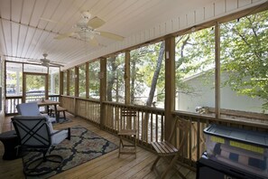 Eine überdachte Veranda mit Holzboden, Deckenventilator und großen Fenstern mit Blick auf Bäume und ein Haus.