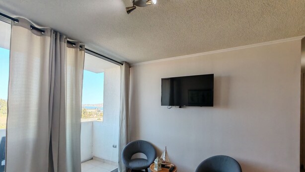 A room with a TV mounted on the wall, two chairs, a small table with a model ship, and a view through a window.