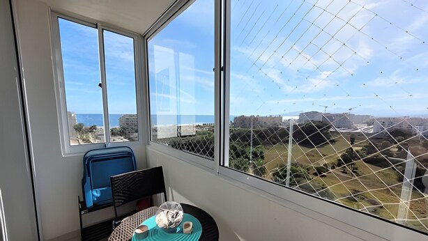 Un balcón con una mesa, una silla y vistas a un paisaje costero con edificios y una fortaleza.