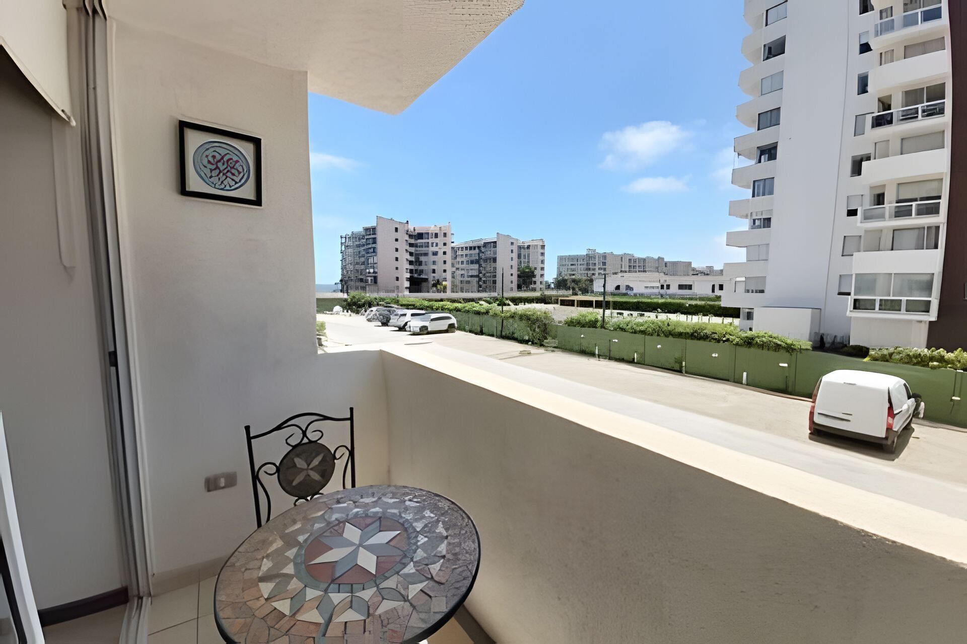 Un balcon avec une table et une chaise carrelées, un tableau encadré et une vue sur un immeuble à plusieurs étages, avec des voitures garées et une clôture verte.