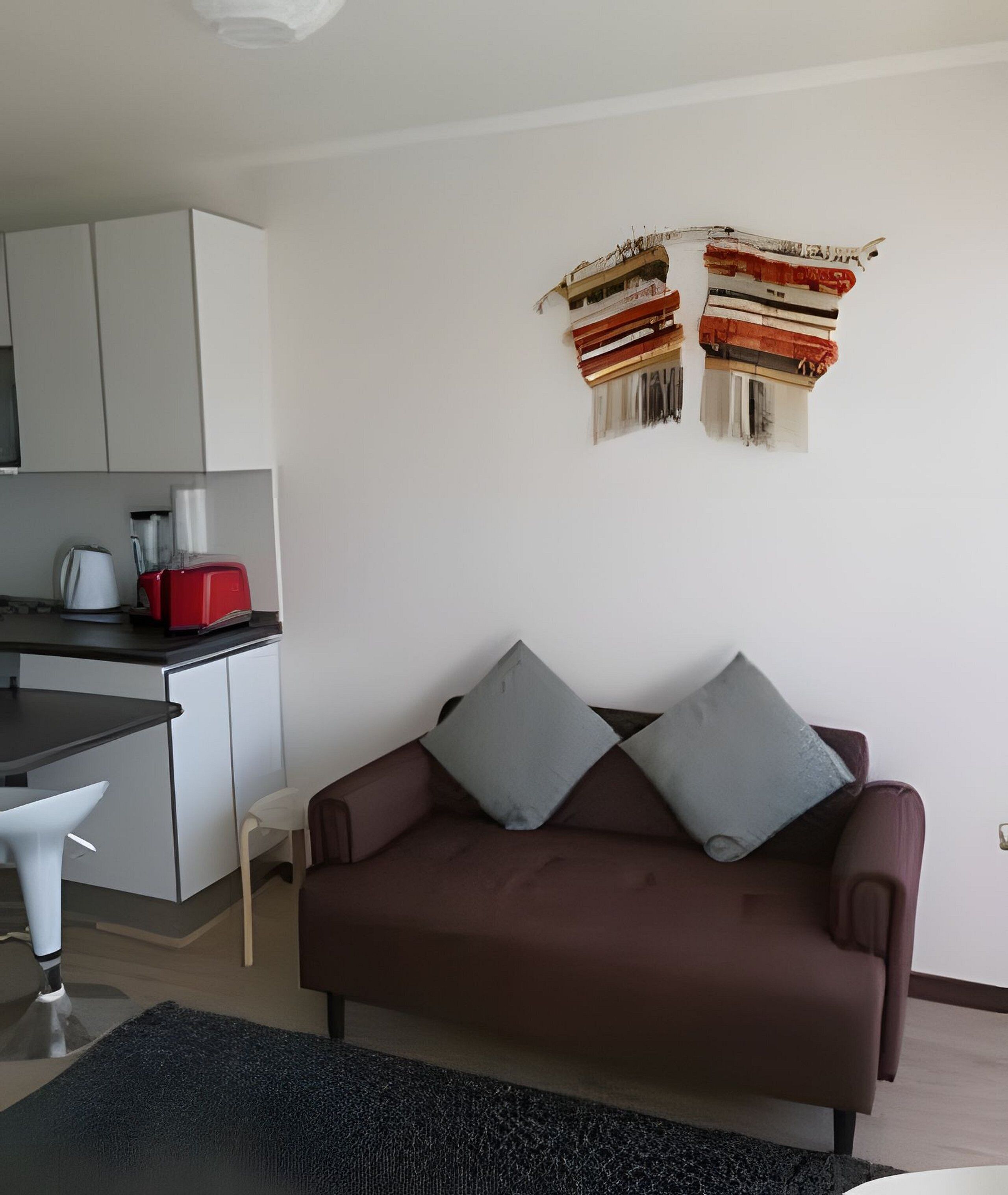 Un salon avec un canapé marron, deux coussins gris, une chaise blanche, un grille-pain rouge, une bouilloire blanche et un meuble blanc.