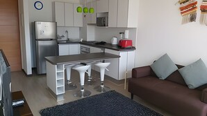 A modern kitchen with white cabinets, a refrigerator, a microwave, and a red toaster. There is a brown sofa with two cushions and a black rug.
