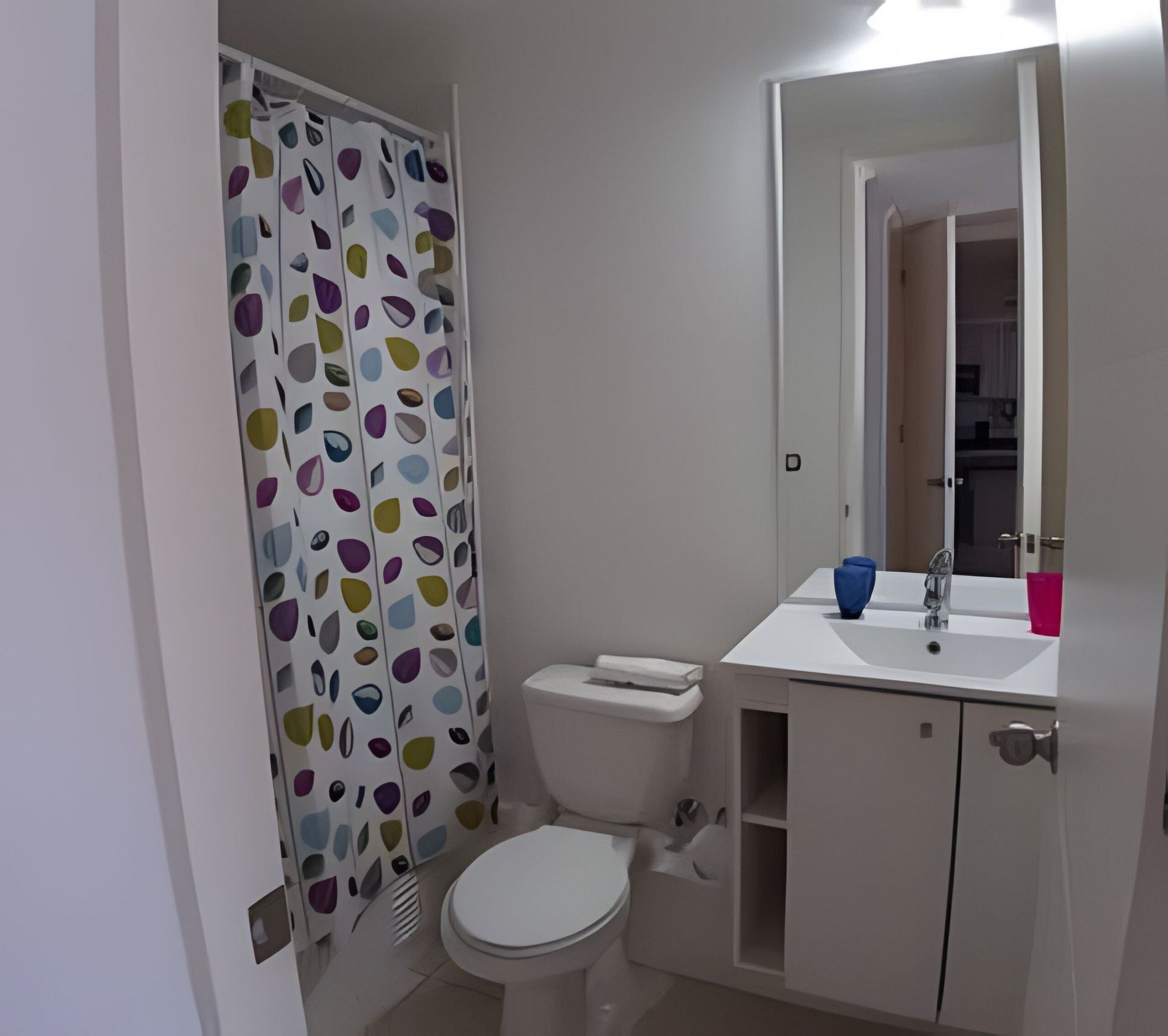 Un baño con inodoro, lavabo, espejo y cortina de ducha con un estampado colorido.