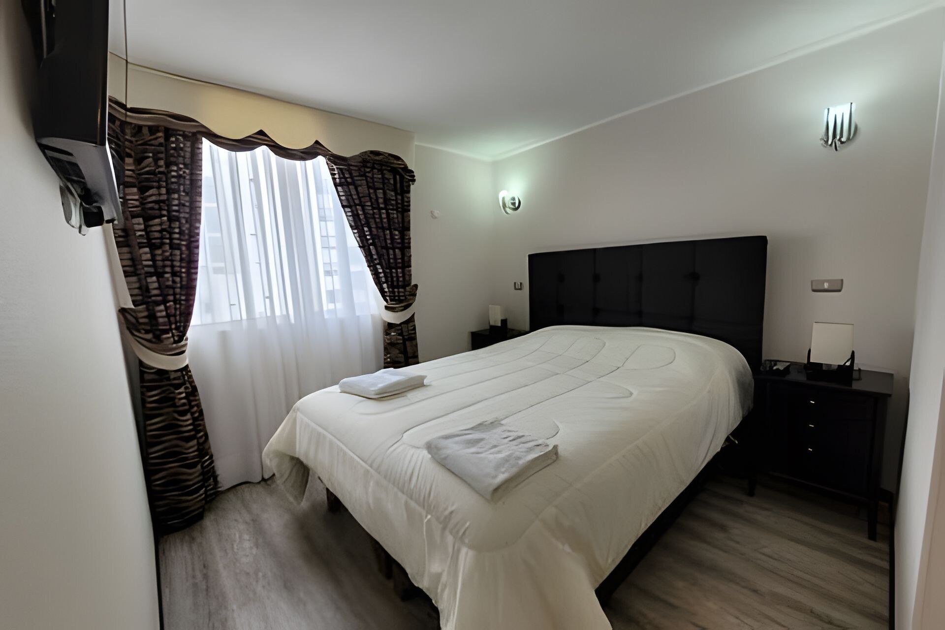 Een slaapkamer met een bed, een nachtkastje, een wandlamp en een raam met gordijnen.