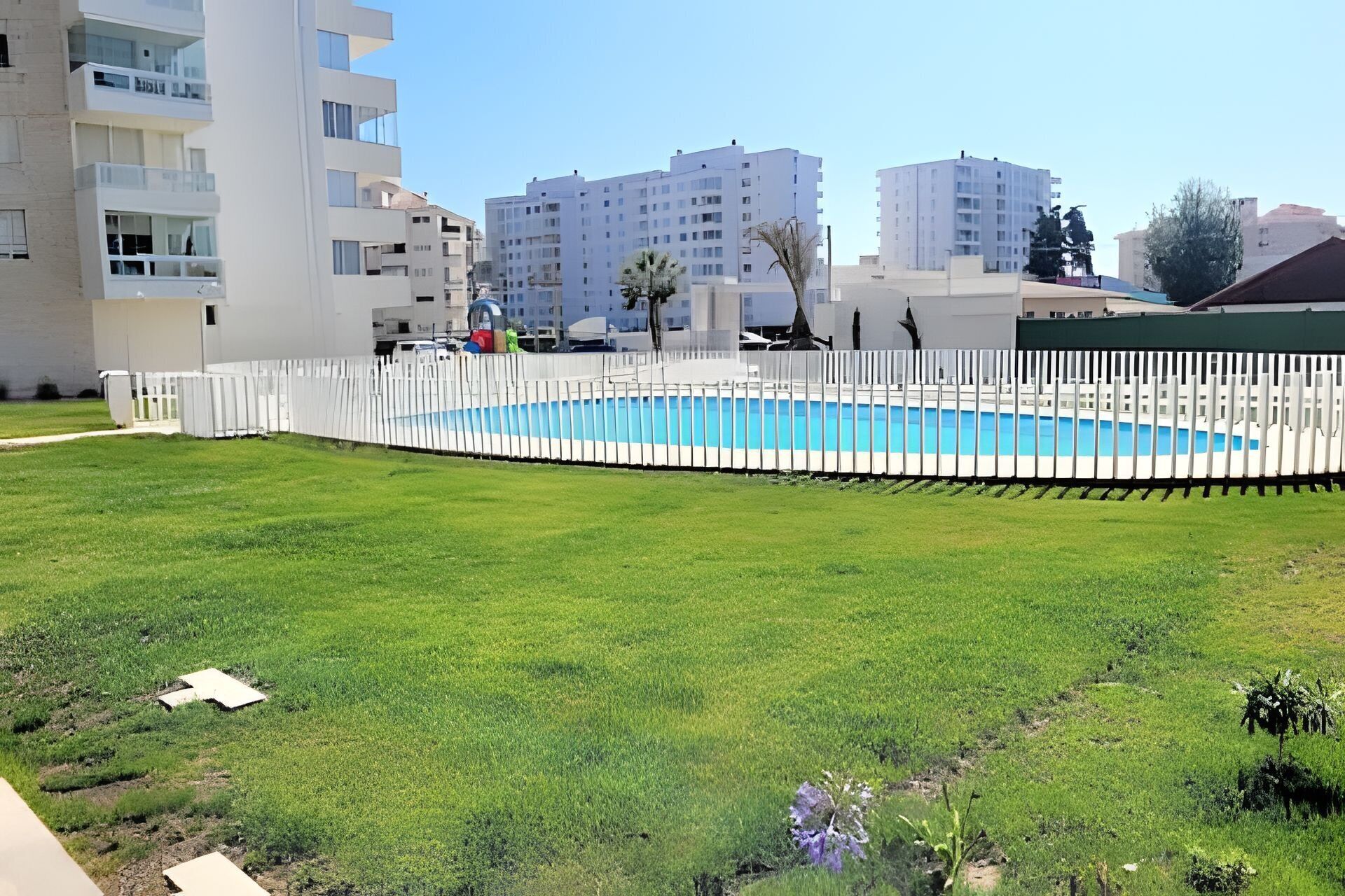 Una piscina rodeada por una cerca blanca, con un césped al frente y edificios residenciales al fondo.