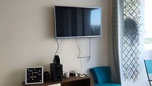 Un salon moderne avec un téléviseur à écran plat fixé au mur, un meuble de rangement en bois doté de tiroirs, un fauteuil bleu et une table ronde agrémentée d’un objet décoratif.