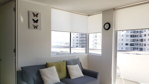 Ett modernt vardagsrum med en grå soffa, en klocka och stora fönster med utsikt över en byggnad.