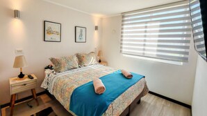 A bedroom with a bed, two wall-mounted lamps, a bedside table with a lamp, a window with blinds, and two framed pictures on the wall. - Dept. 5th Floor, Pacífico 2800 (La Serena)