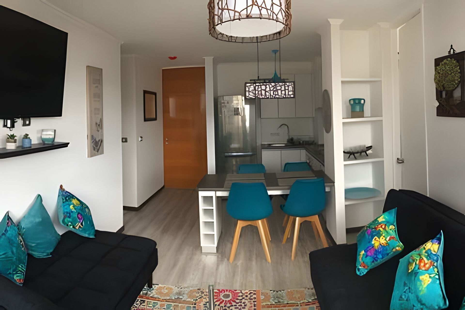 Un salon moderne avec un canapé noir, des coussins turquoise, un tapis à motifs et un coin cuisine comprenant une table à manger et des chaises.