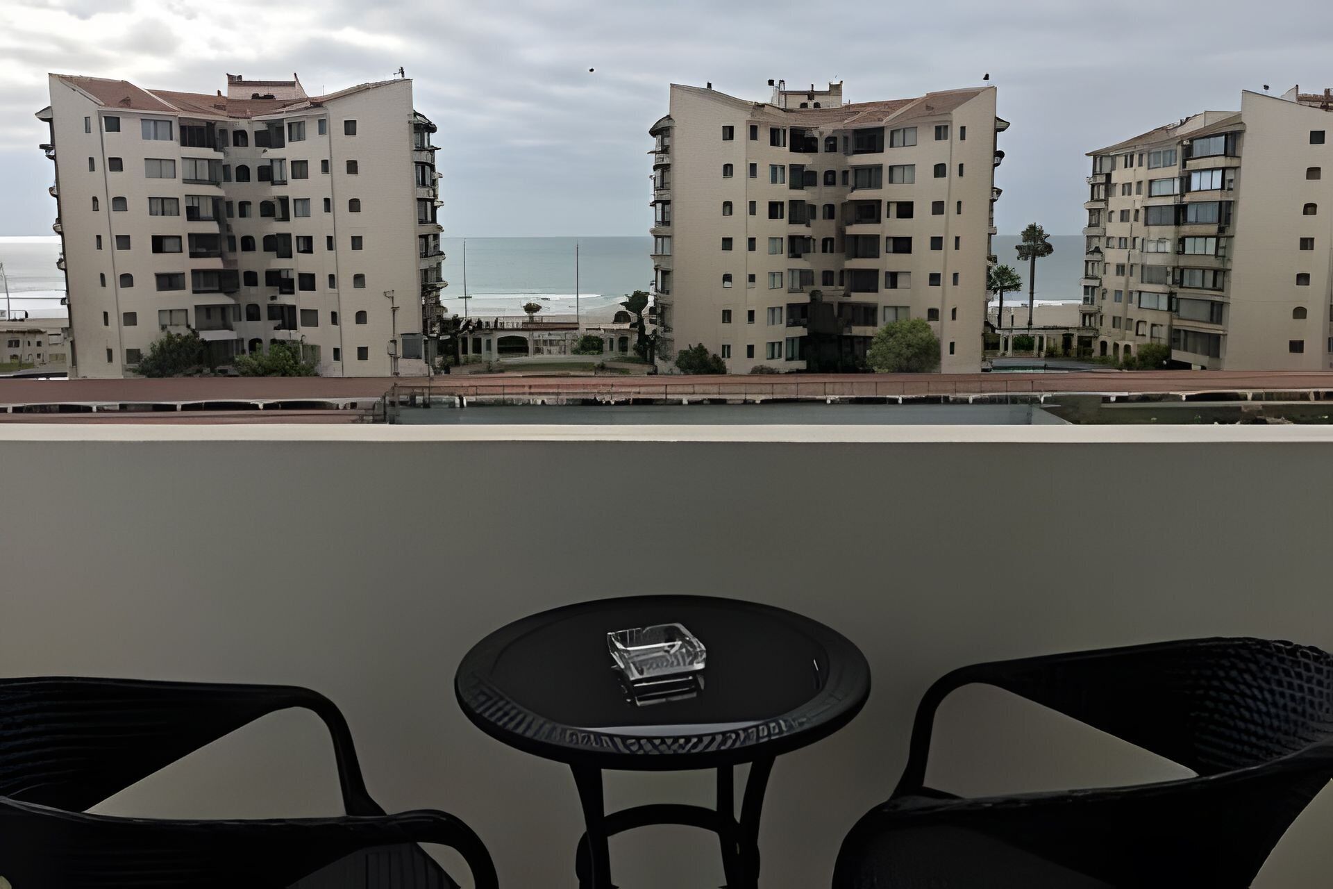 A balcony with two chairs and a small table, overlooking a coastal area with buildings and a view of the sea.