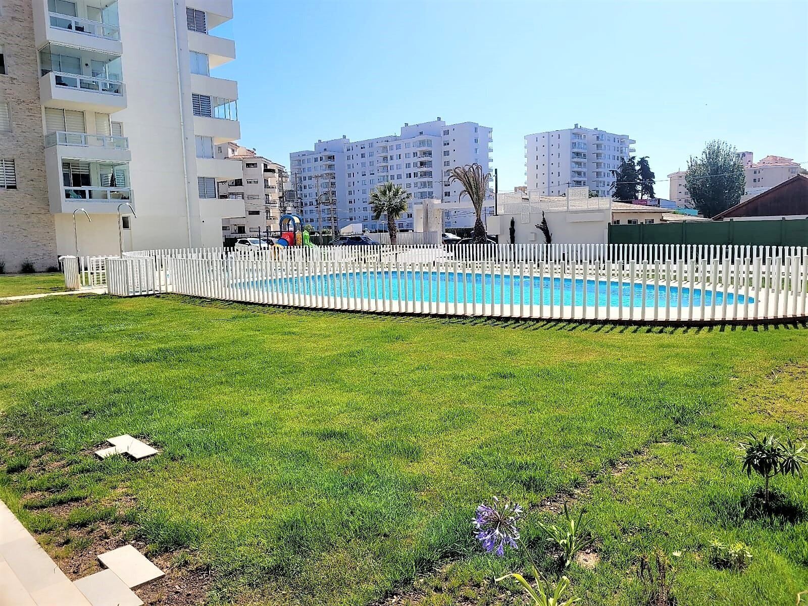 Una piscina rodeada por una valla blanca, con un área verde en primer plano y edificios de varios pisos al fondo.