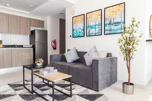 A modern living room with a grey sofa, a coffee table, and a bookshelf. There is a kitchen area with wooden cabinets and a stainless steel refrigerator. - Bloomfields Studio in The View Raha Beach (Abu Dhabi)
