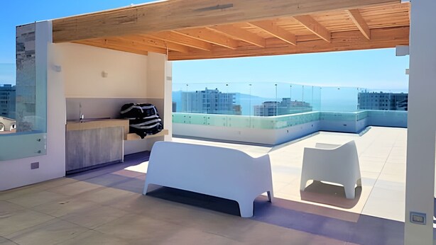 Eine Dachterrasse mit Holzboden, einer weißen Bank, einem kleinen weißen Tisch und einem Glasgeländer mit Blick auf eine Stadtlandschaft.