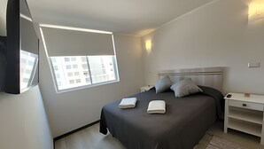 Ein modernes Hotelzimmer mit einem großen Bett, einem Nachttisch, einem kleinen Tisch daneben, einem an der Wand befestigten Fernseher und einem Fenster mit Blick auf Gebäude.