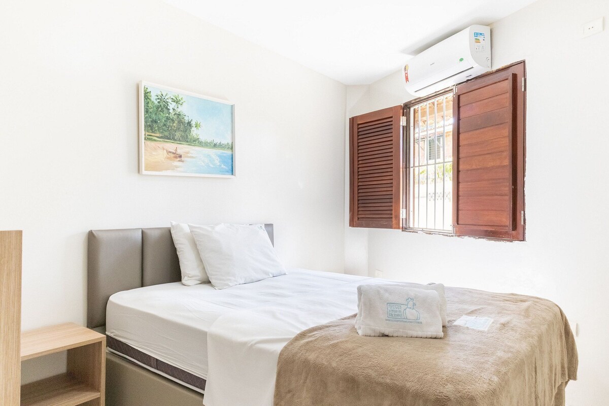 Ein Einzelbett mit Kopfteil, ein Nachttisch aus Holz, ein Handtuch, ein Gemälde mit Strandmotiv und ein Fenster mit Holzjalousien.