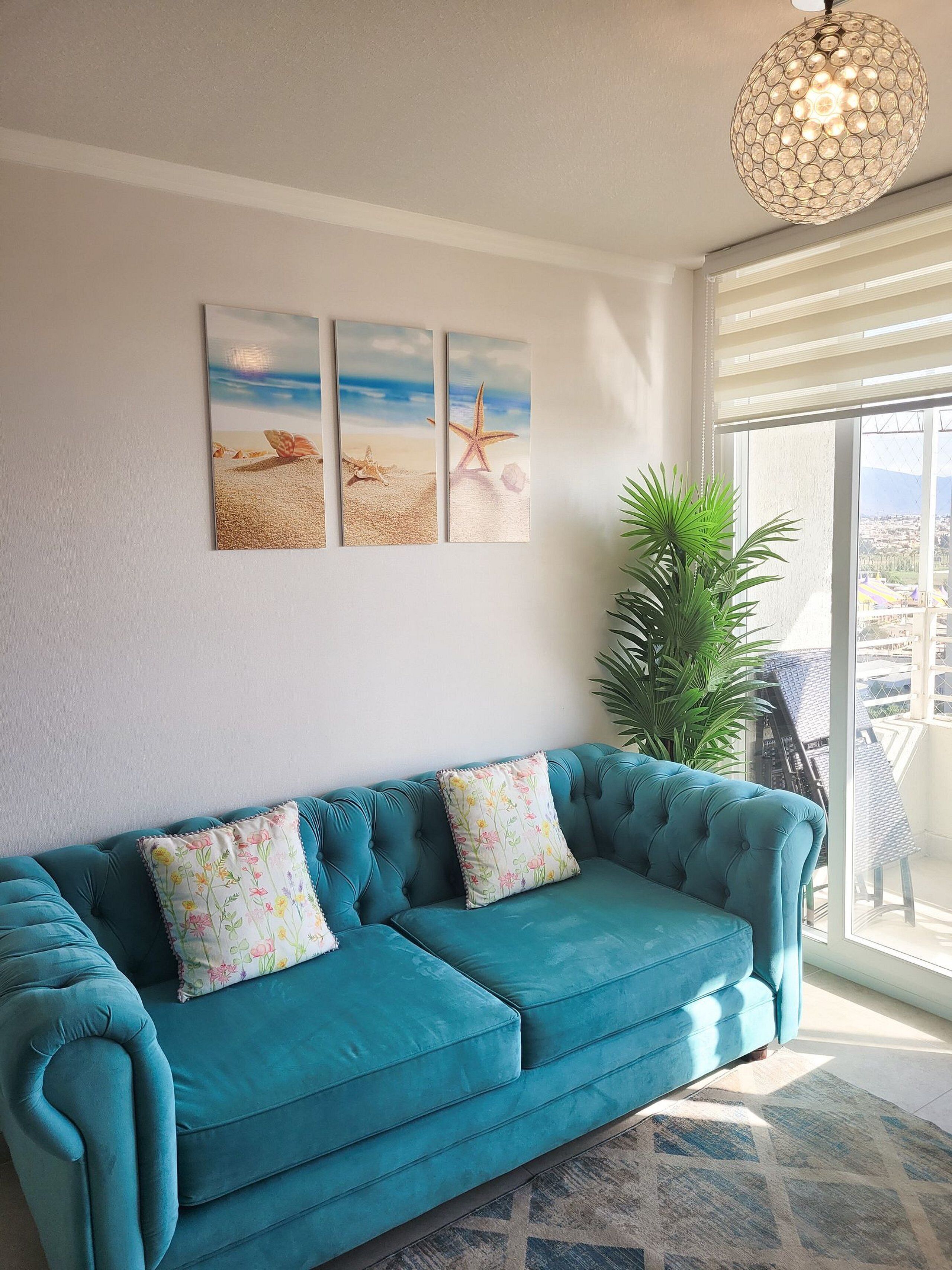 Un canapé de couleur turquoise agrémenté de coussins à motifs floraux, une œuvre d’art représentant une étoile de mer accrochée au mur, et une grande fenêtre offrant une vue imprenable.