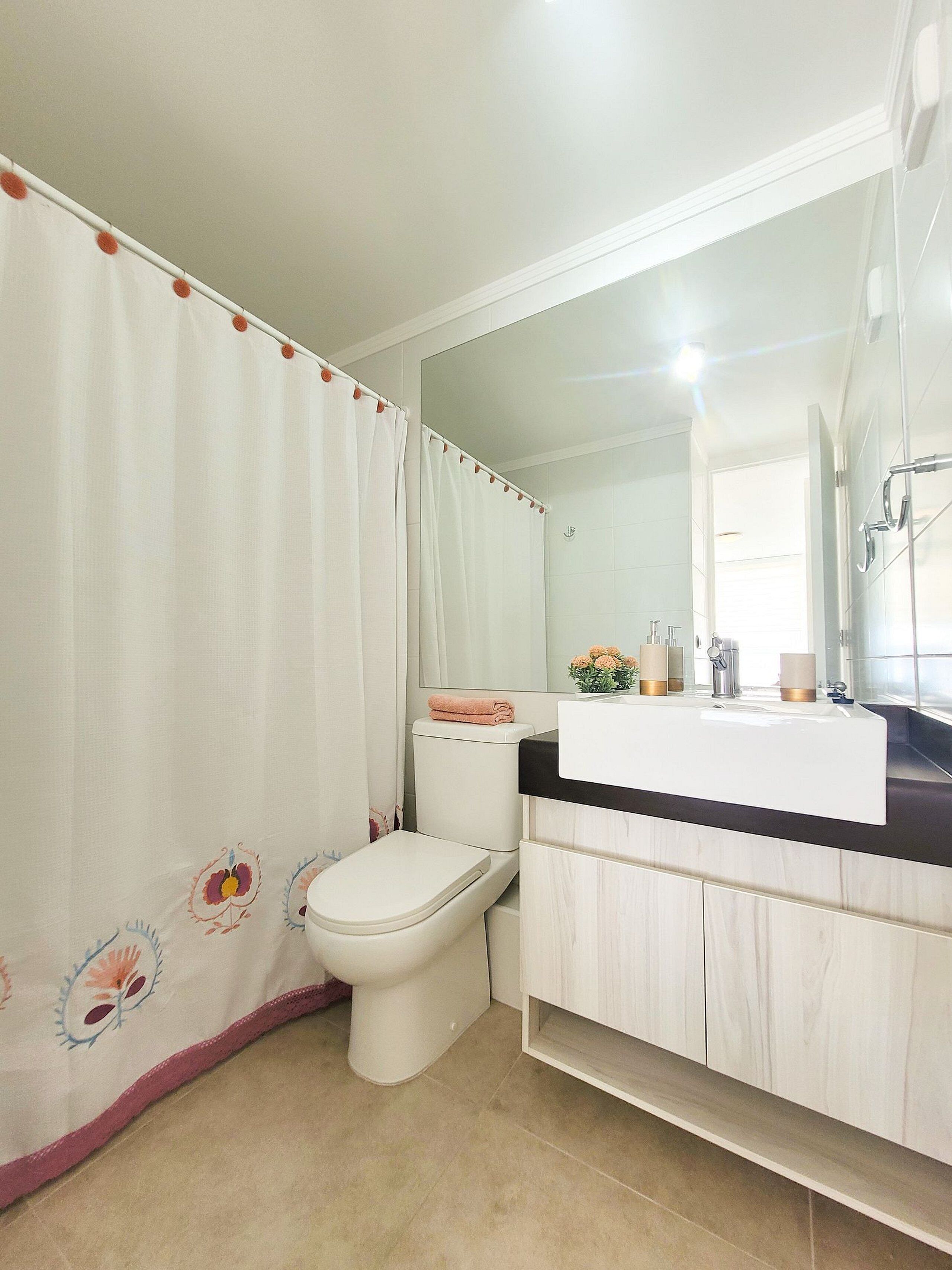 Un baño con inodoro, lavabo con espejo y cortina de ducha estampada con motivos florales.