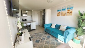 A modern living room with a blue sofa, a white coffee table, and a kitchen area with a refrigerator and a TV. - Dept. 14th Floor, Marina Costa (Coquimbo)
