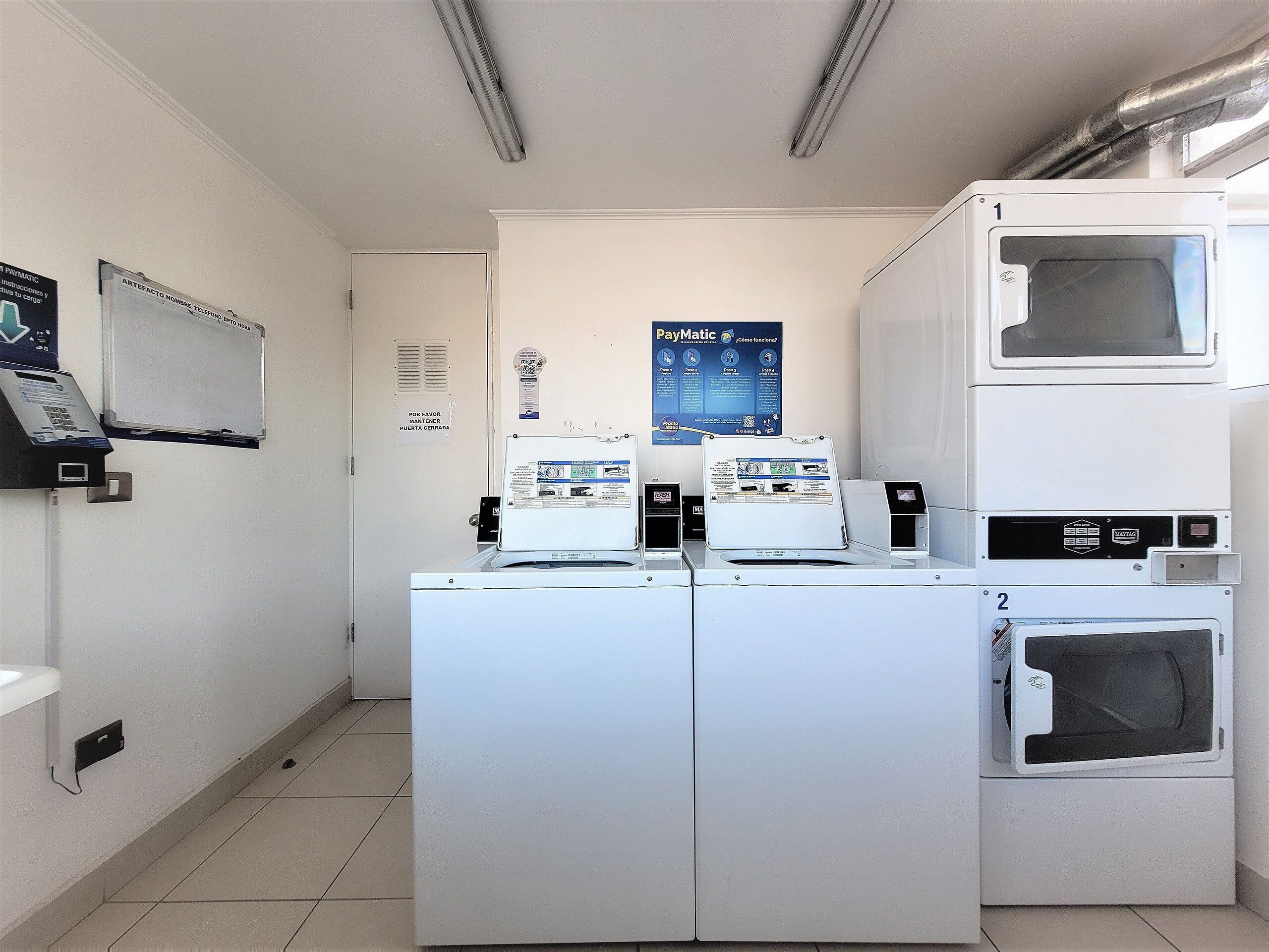 A laundry facility with two washing machines, a dryer, a Paymatic self-service kiosk, and a whiteboard.
