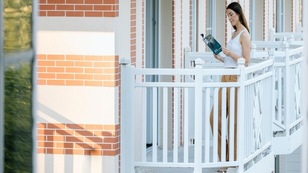 Una persona está de pie en un balcón con barandilla blanca, leyendo un libro.
