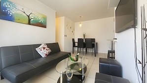 Una sala de estar moderna con un sofá, una mesa de centro de vidrio, un comedor con sillas y un televisor de pantalla plana montado en la pared.