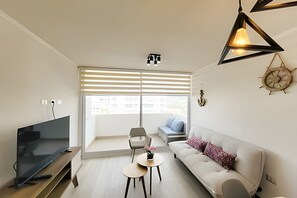 A modern living room with a flat-screen TV, a sofa, a coffee table, and a large window with blinds. - Dept. 5th Floor, Distrito Verde (La Serena)