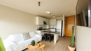 A modern living room with a sofa, a small round table with chairs, a kitchen area with a dining table, and a potted plant. - Dept. 10th Floor, Cond. Pacific II (La Serena)