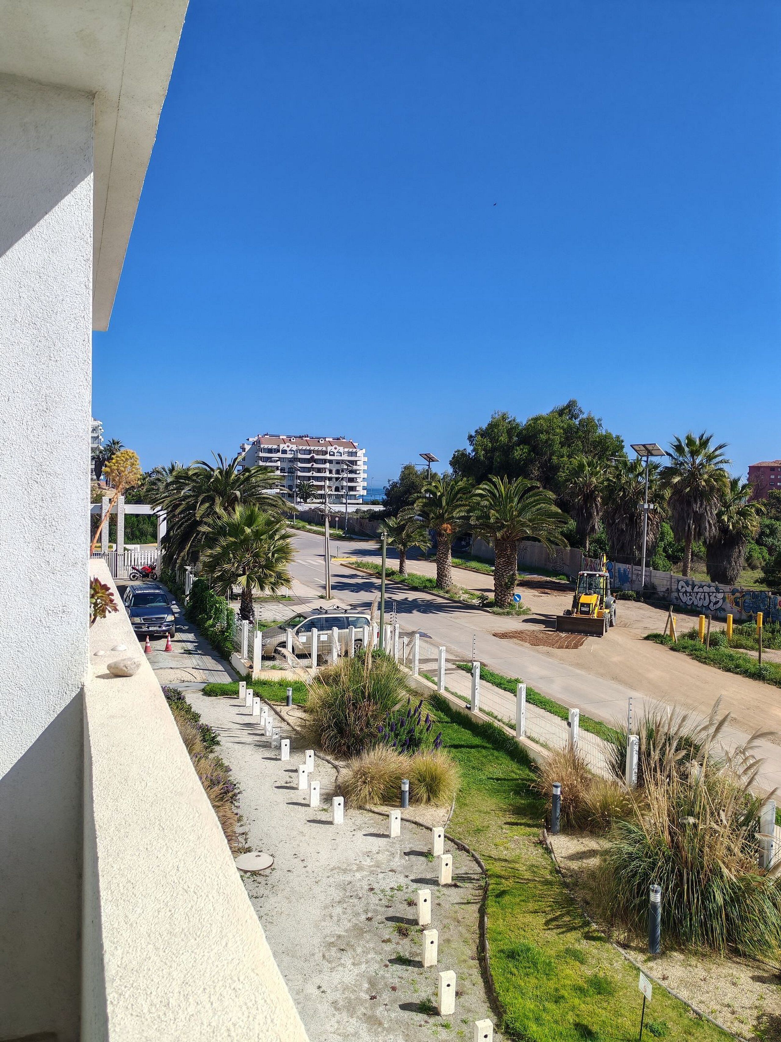 Un balcón con vista a una calle con coches estacionados, un vehículo de construcción y un edificio al fondo.