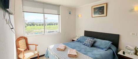 A bedroom with a bed, a chair, a bedside table, a nightstand, a window, and a painting on the wall.