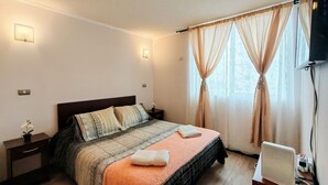 Ein ordentlich bezogenes Bett mit einem gemusterten Überwurf, ein Nachttisch mit Lampe und ein Fenster mit durchsichtigen Vorhängen.