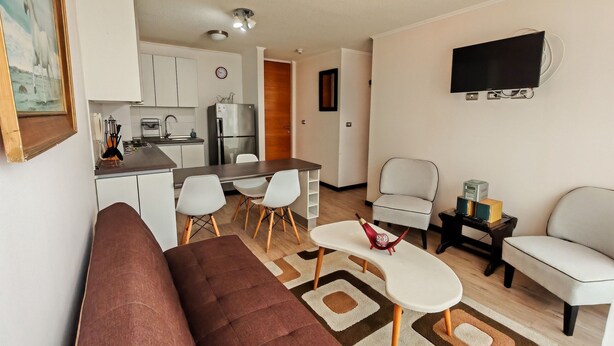 Ein modernes Wohnzimmer mit einem braunen Sofa, weißen Stühlen, einem kleinen weißen Tisch und einem an der Wand montierten Fernseher.