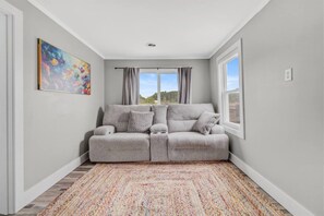 A room with a grey sofa, patterned cushions, a colorful rug, a painting on the wall, and a window with curtains.